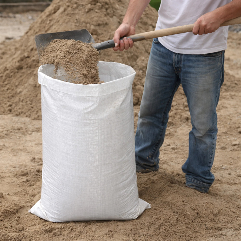 Person filling a large white bag with sand using a shovel.